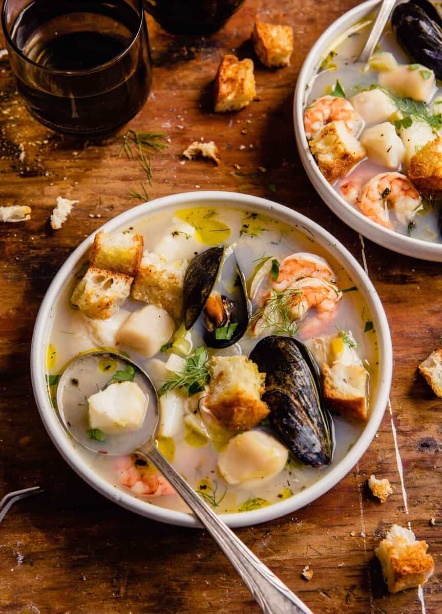 mussels, shrimp, scallops and chunks of white fish in broth with herbs and croutons in a white bowl set on a wood table