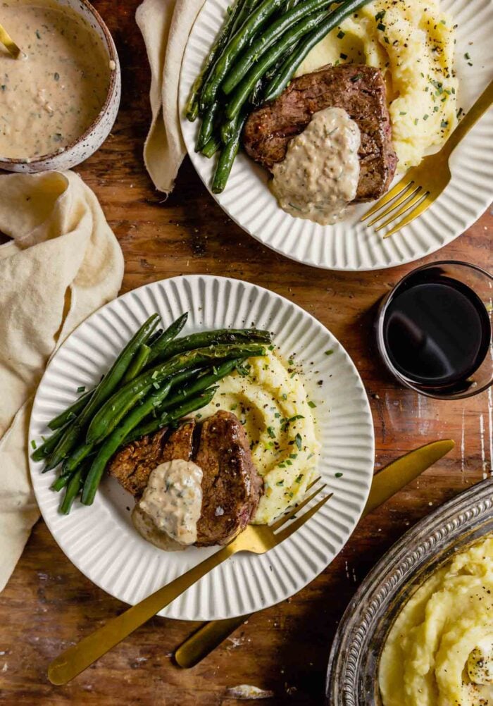 white plates set on a wood table topped with mashed potatoes, pan seared filet mignon, and green beans