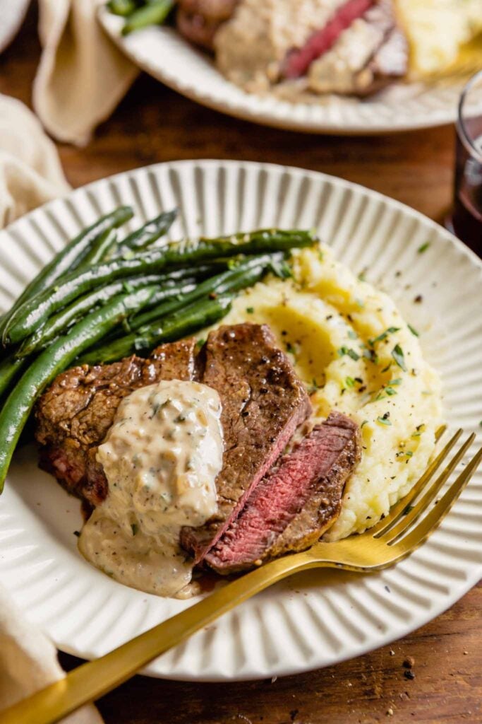 white plates set on a wood table topped with mashed potatoes, pan seared filet mignon, and green beans