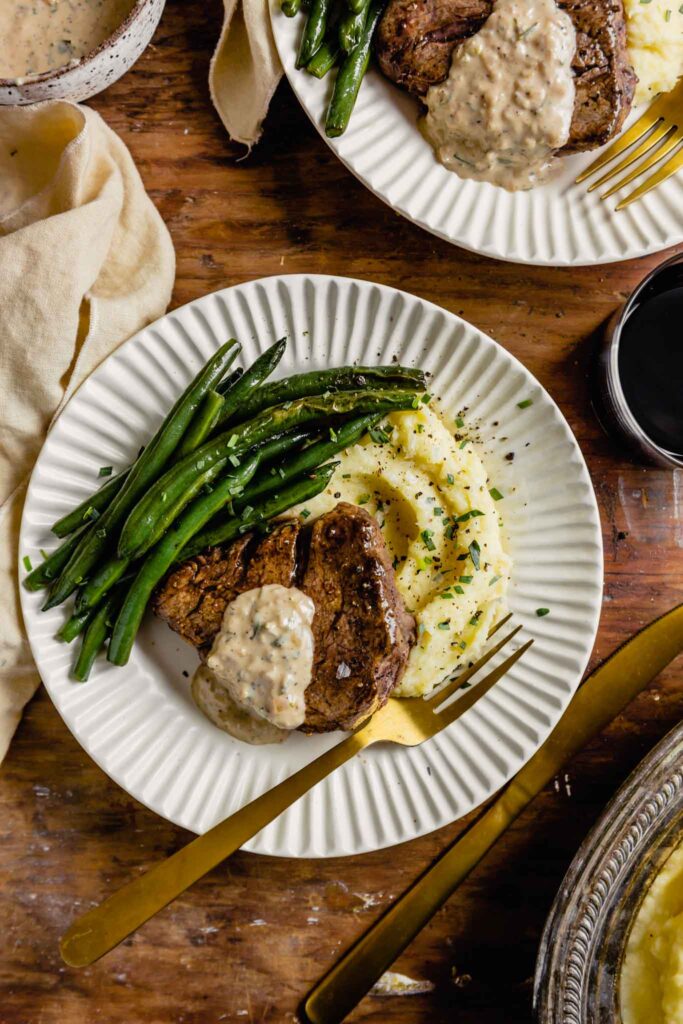 white plates set on a wood table topped with mashed potatoes, pan seared filet mignon, and green beans