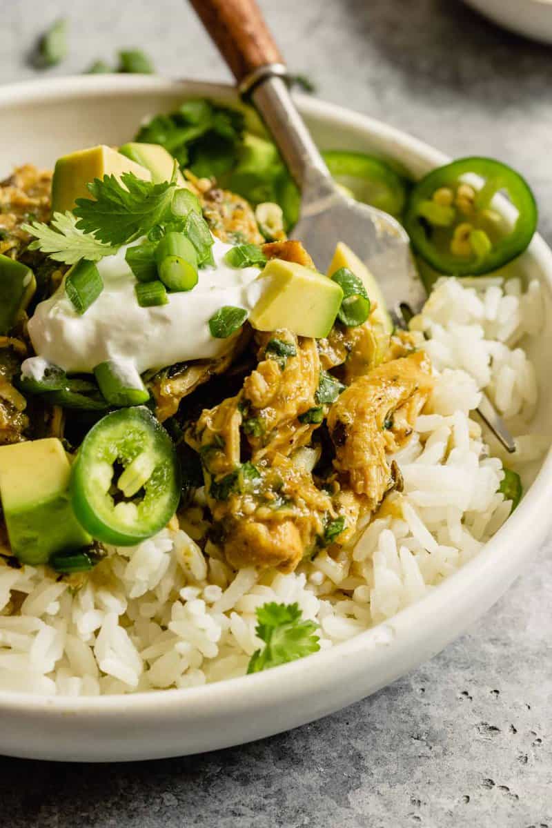 close up of salsa verde chicken on top of white rice in a white bowl topped with avocado, jalapeno slices and cilantro