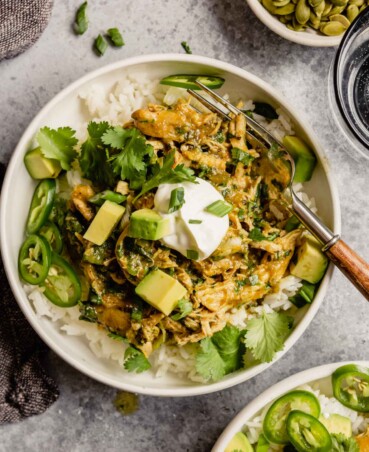 shredded salsa verde chicken in top of white rice in a white bowl garnished with sour cream, avocado, scallions, and cilantro
