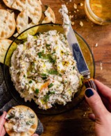 creamy white dip in a green glass bowl with a pair of hands spreading some dip on a cracker