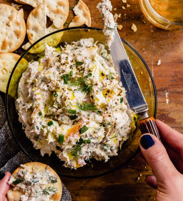 creamy white dip in a green glass bowl with a pair of hands spreading some dip on a cracker