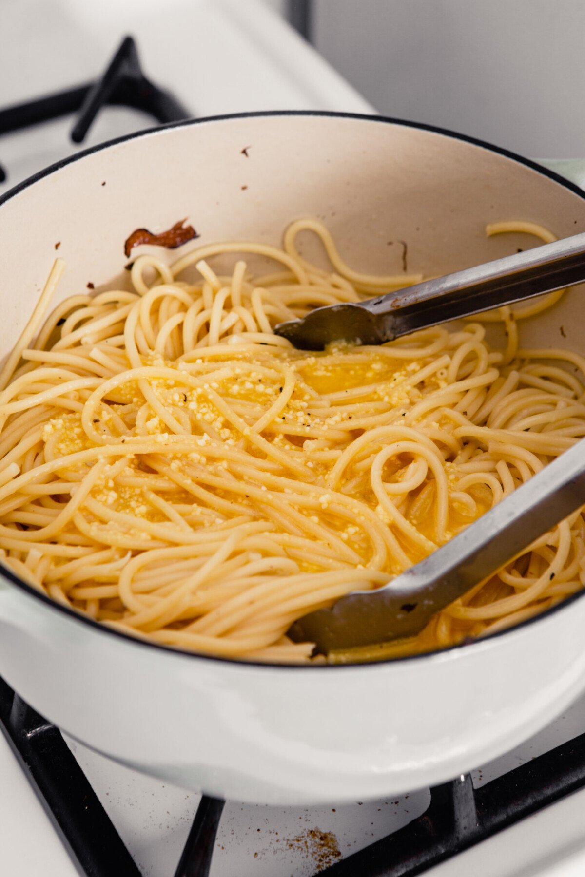 A pair of tongs set in a Dutch oven full of cooked pasta with an egg and cheese mixture poured over top. 