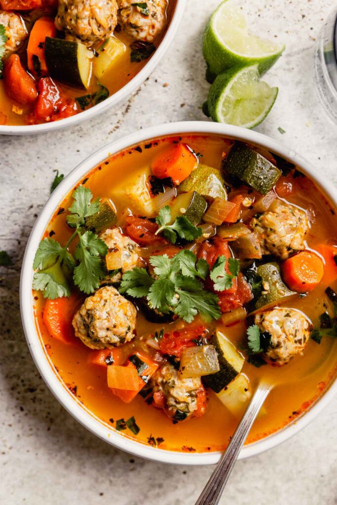 brothy vegetable soup with meatballs in a white bowl with lime wedges set around