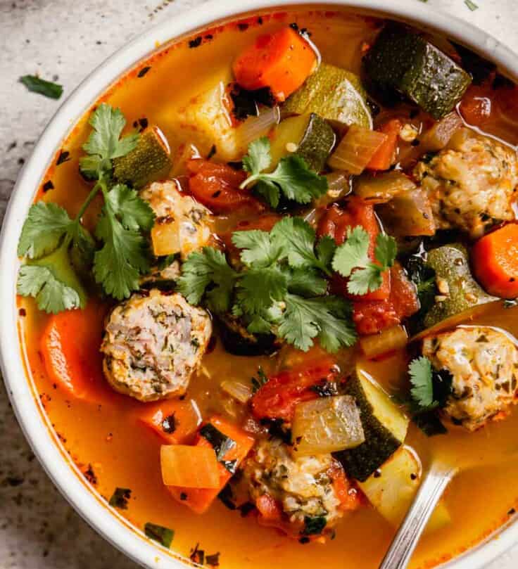 brothy vegetable soup with meatballs in a white bowl with lime wedges set around