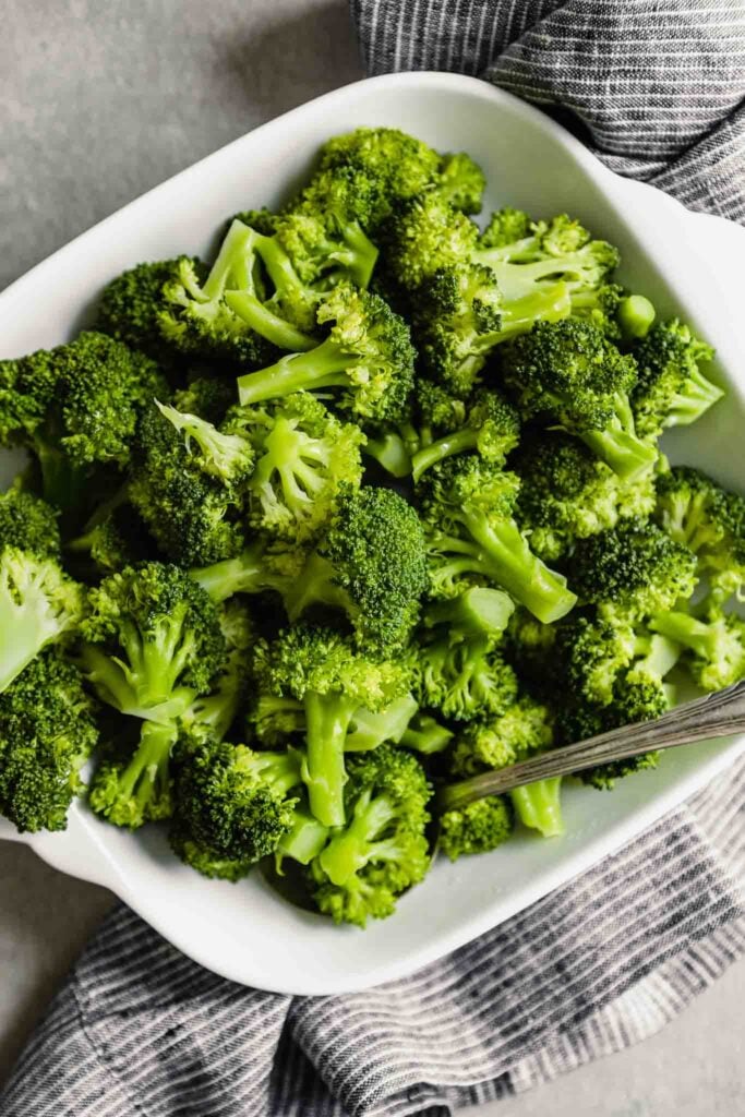 steamed broccoli florets in a white square bowl set on a gray surface with a stripped blue linen napkin