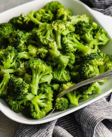 steamed broccoli florets in a white square bowl set on a gray surface with a stripped blue linen napkin