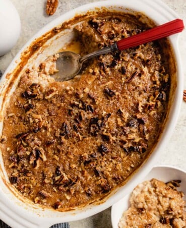 baking dish filled with a sugary baked oatmeal with a red handled spoon set in the dish and oatmeal in a small white bowl off to the side