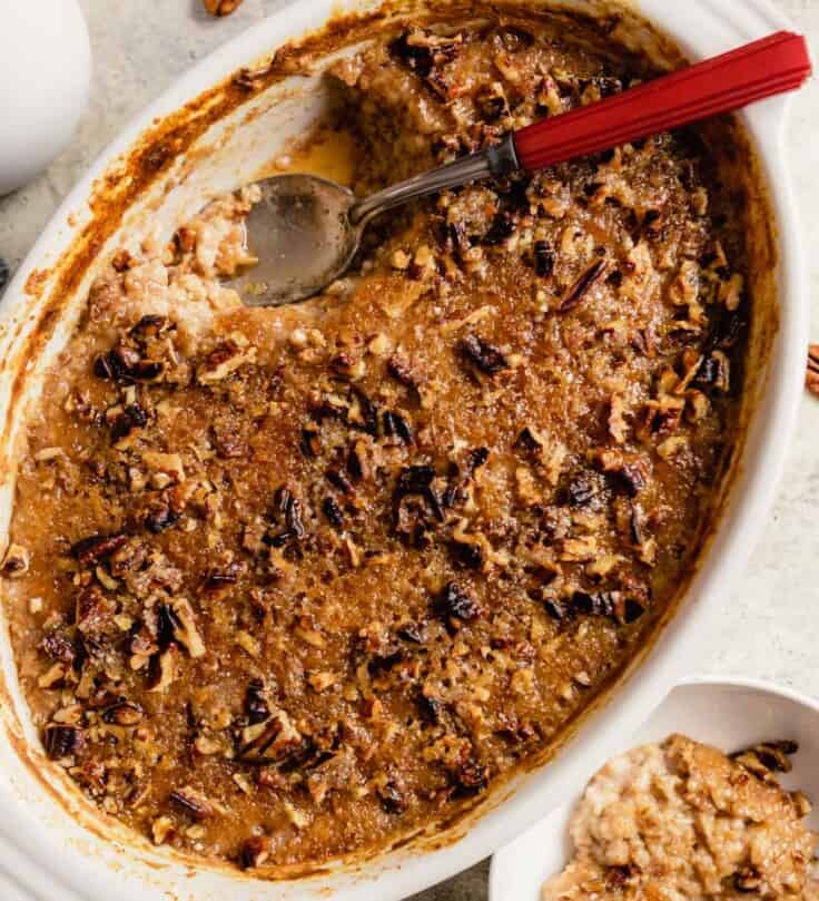 baking dish filled with a sugary baked oatmeal with a red handled spoon set in the dish and oatmeal in a small white bowl off to the side