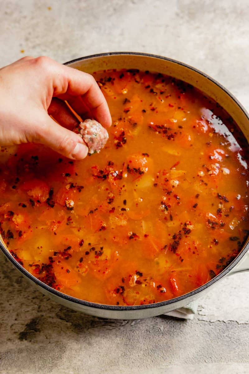 a hand dropping a meatball into a pot of simmering broth