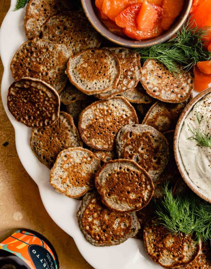 small buckwheat pancakes stacked on a white platter with smoked salmon, orange segments, and cream cheese