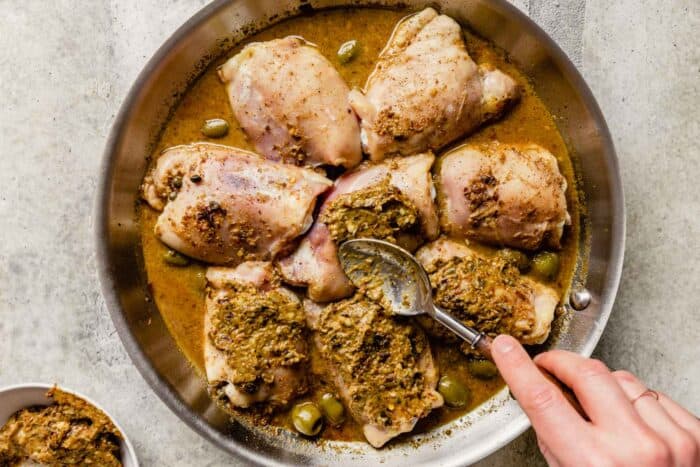 olive paste being spread over chicken thighs in a skillet with broth and olives