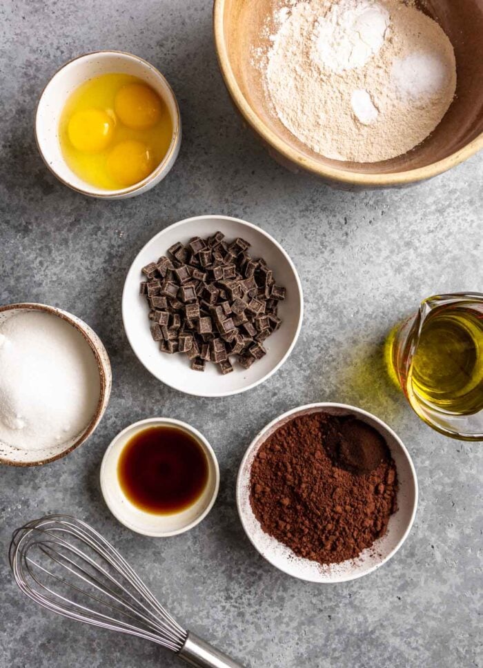 flour, cocoa powder, chocolate chips, olive oil, sugar, eggs, vanilla and a whisk set on a blue-gray counter