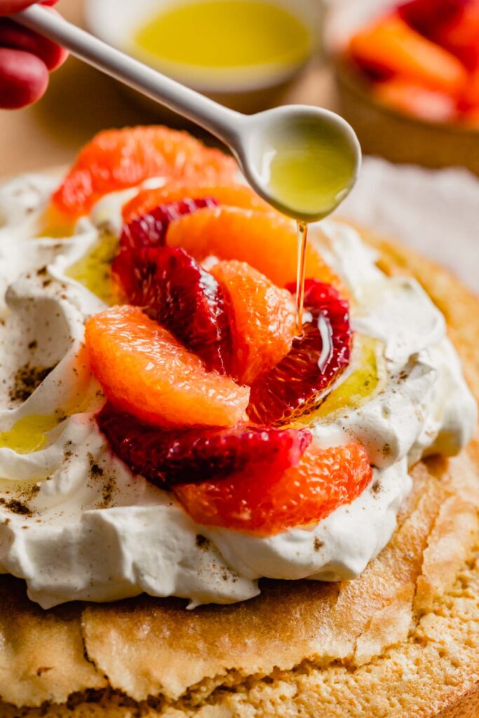 olive oil being drizzled from a small white spoon over oranges and whipped cream on top of a cake