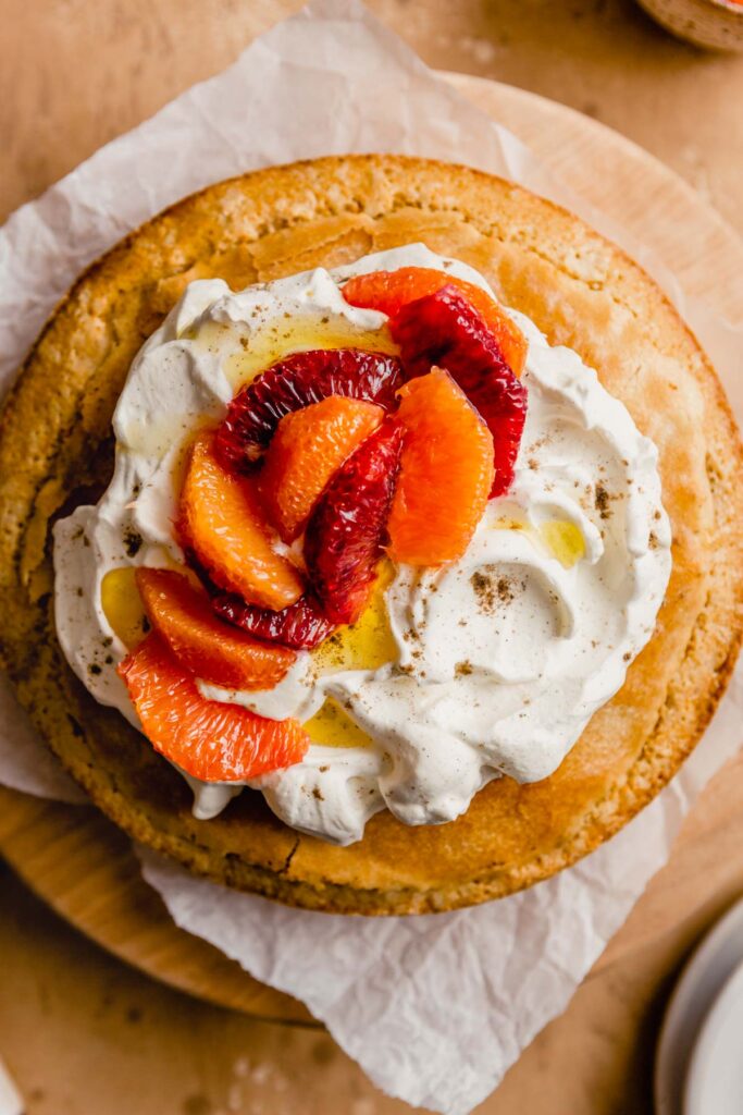 a whole round cake topped with whipped cream and oranges set on a parchment paper on a round wood board 