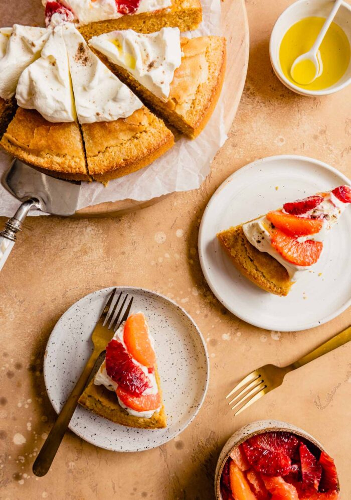 slices of cake topped with whipped cream and oranges on white plates with gold forks, whole round cake cut into pieces set on a wood board off to the top