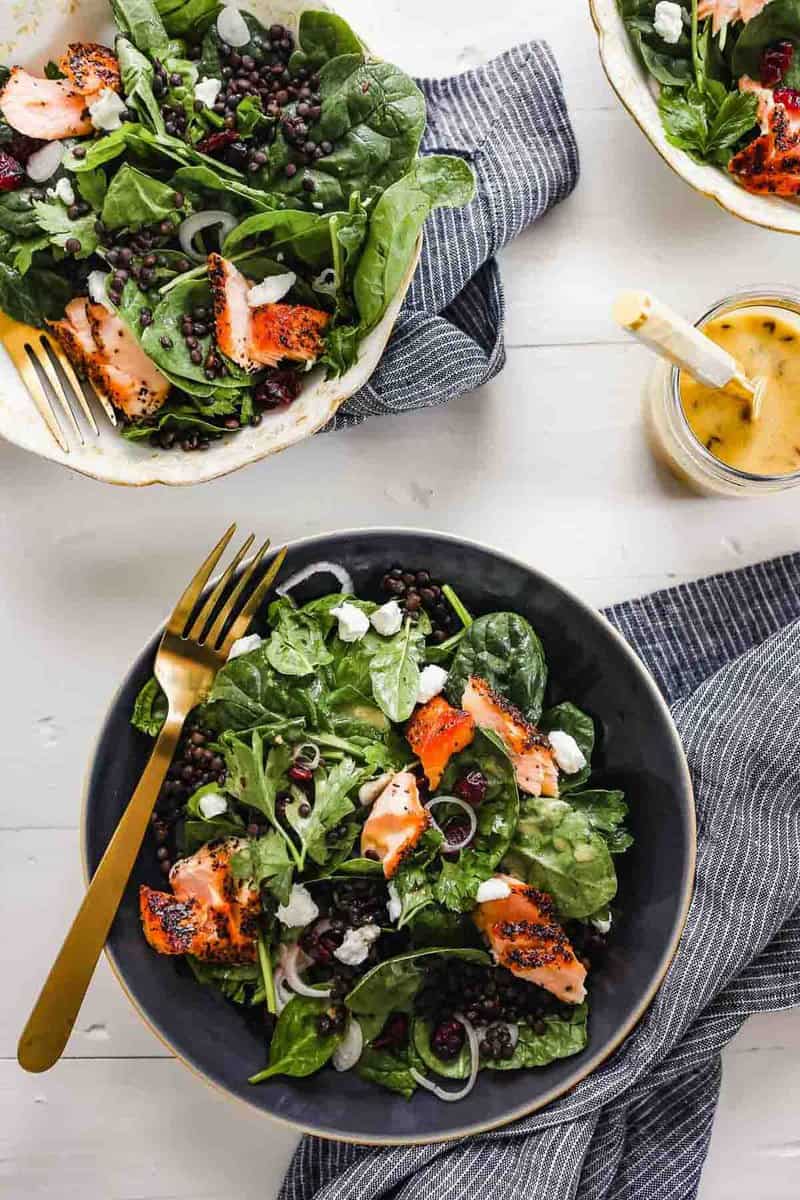 Overhead photograph of lentil and spinach salad with flakes of salmon in a dark blue bowl with a gold fork. Dressing set off to the side in a bowl with a spoon.