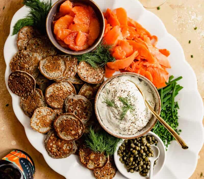 large white platter filled with smoked salmon, mini buckwheat pancakes, dill cream cheese, chives, and caper. Set on an orange table with mimosas and a bottle of sparkling wine