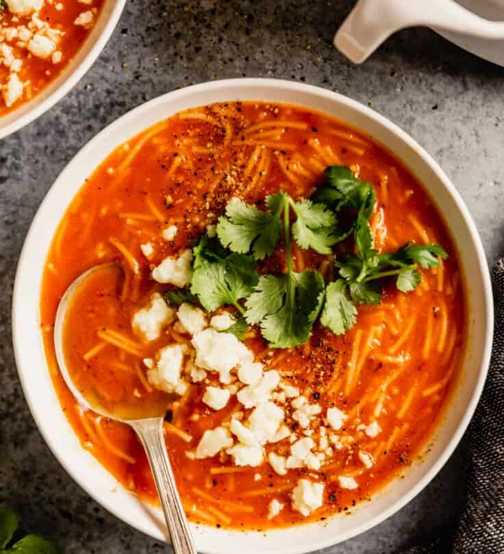 white bowl filled with tomato and noodle soup and topped with cilantro and crumbled cheese