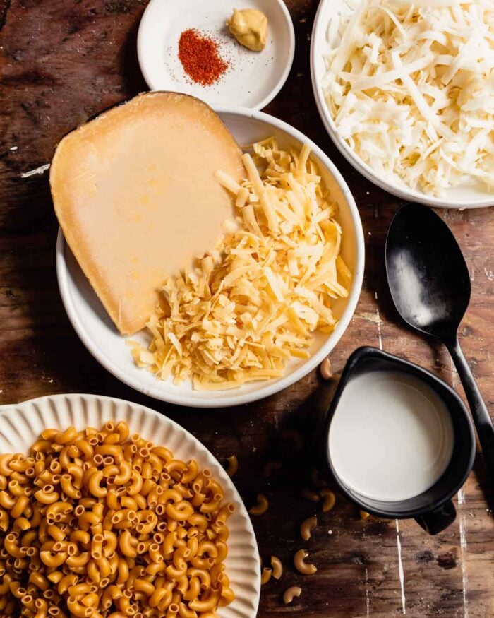 uncooked elbow noodles, shredded cheese, milk, and spices measured out in white bowls on a wood table