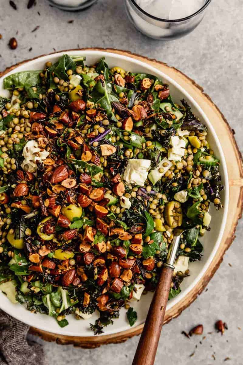 kale and lentil salad in a white bowl set on a wood board topped with feta cheese and toasted almonds