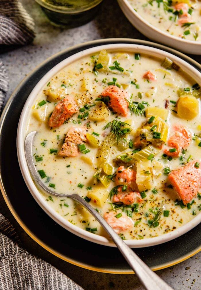white bowl filled with a creamy white soup featuring chunks of salmon. Set on a blue plate with a spoon set in the soup.