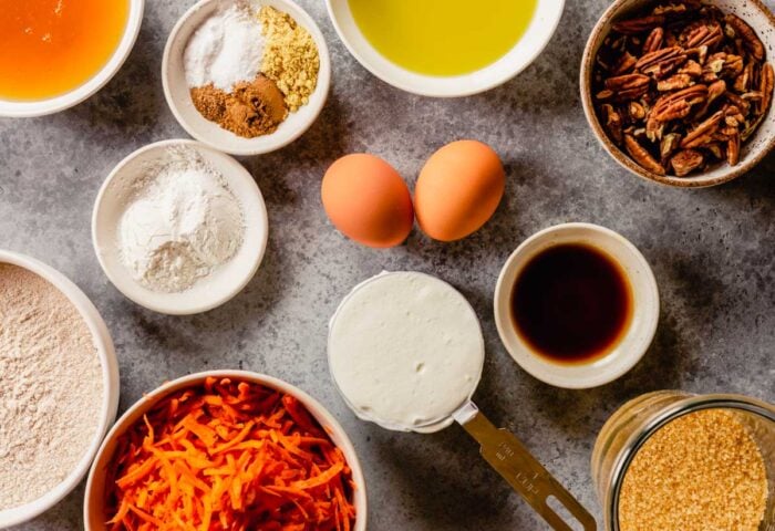 olive oil, honey, shredded carrots, flour, spices, yogurt, and vanilla measured out in bowls. Eggs arranged around bowl