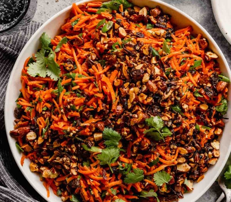 A bowl of carrot raisin salad with nuts, seeds, and fresh cilantro sits on a gray table.