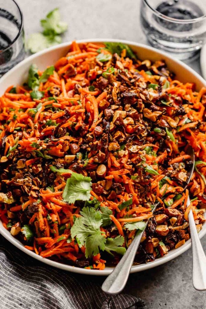shredded carrot salad in a white bowl set on a gray table with glasses set around it