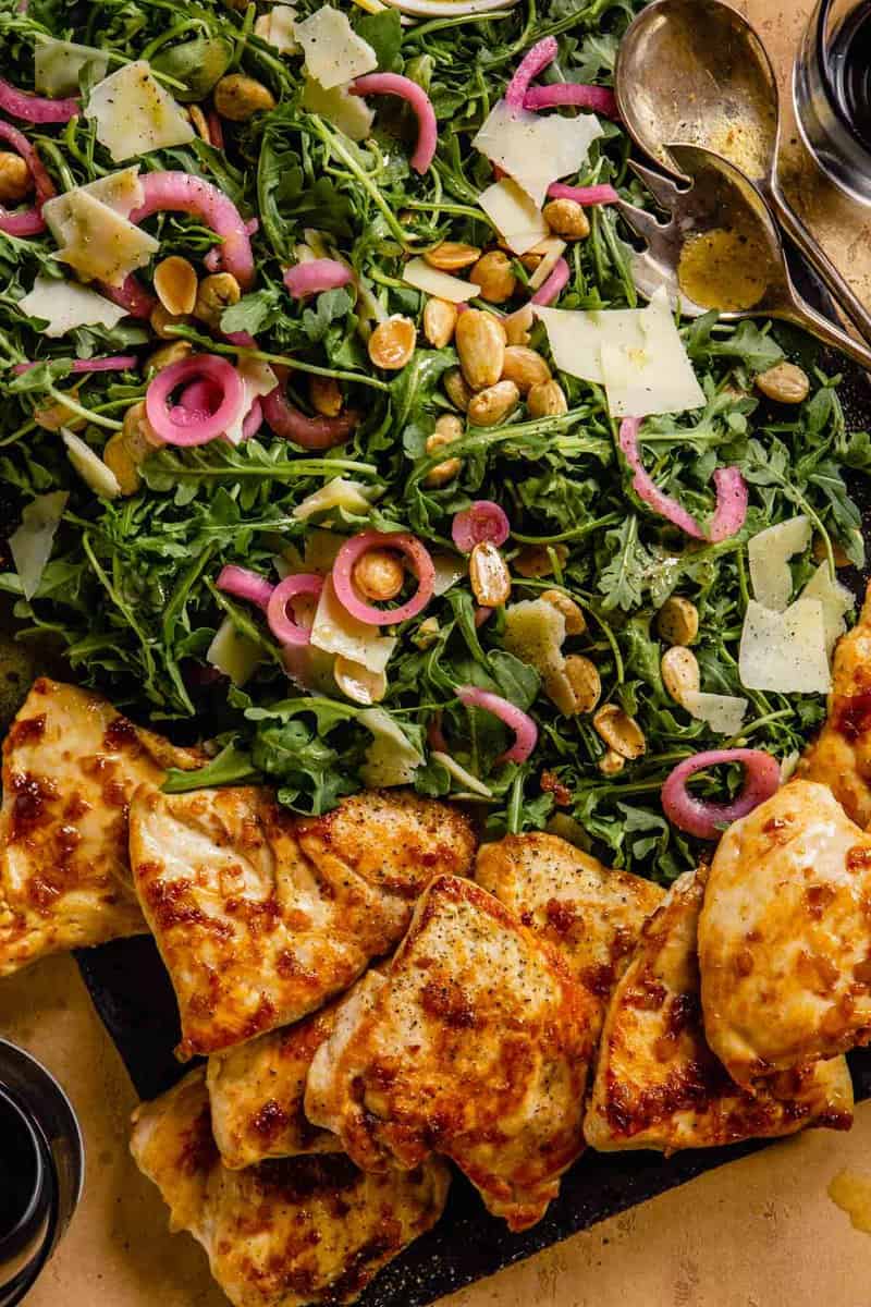 golden brown chicken on a slate board with arugula salad topped with pickled red onion, parmesan and almonds