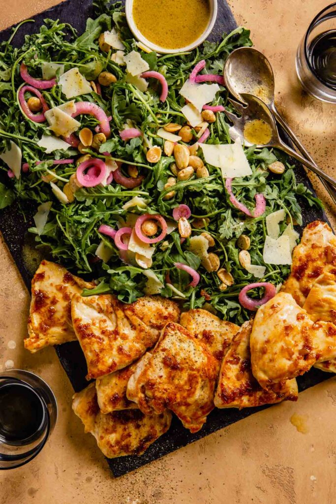golden brown chicken on a slate board with arugula salad topped with pickled red onion, parmesan and almonds