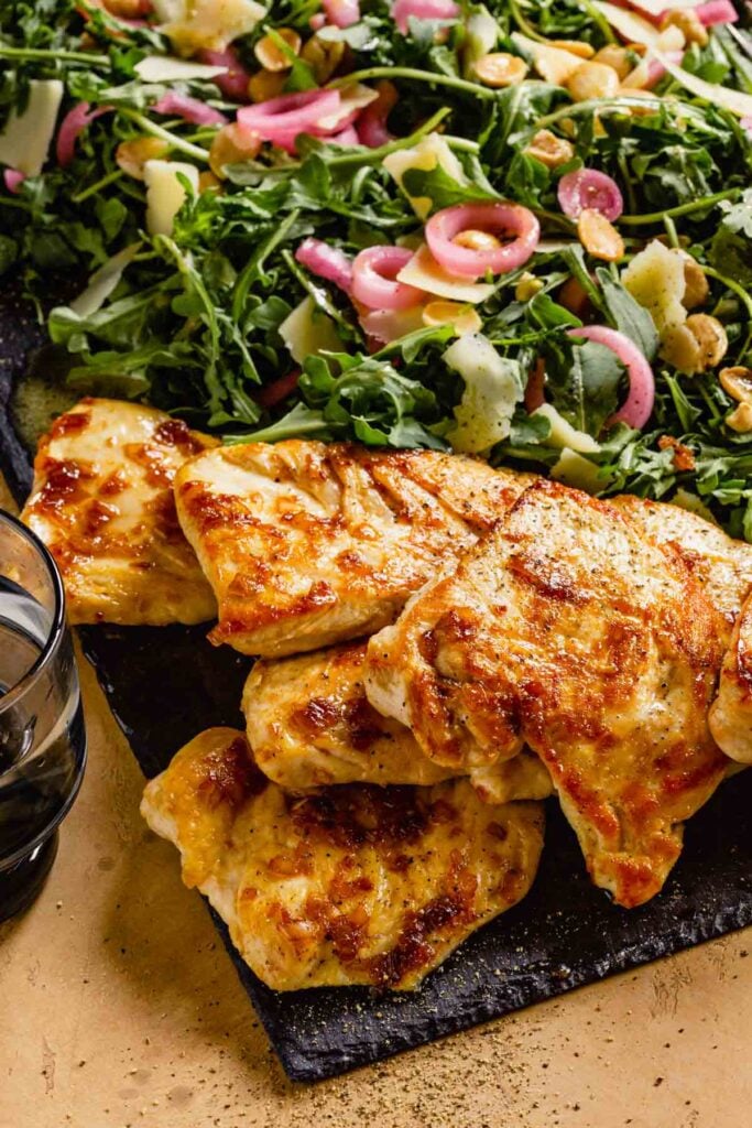 golden brown chicken on a slate board with arugula salad topped with pickled red onion, parmesan and almonds