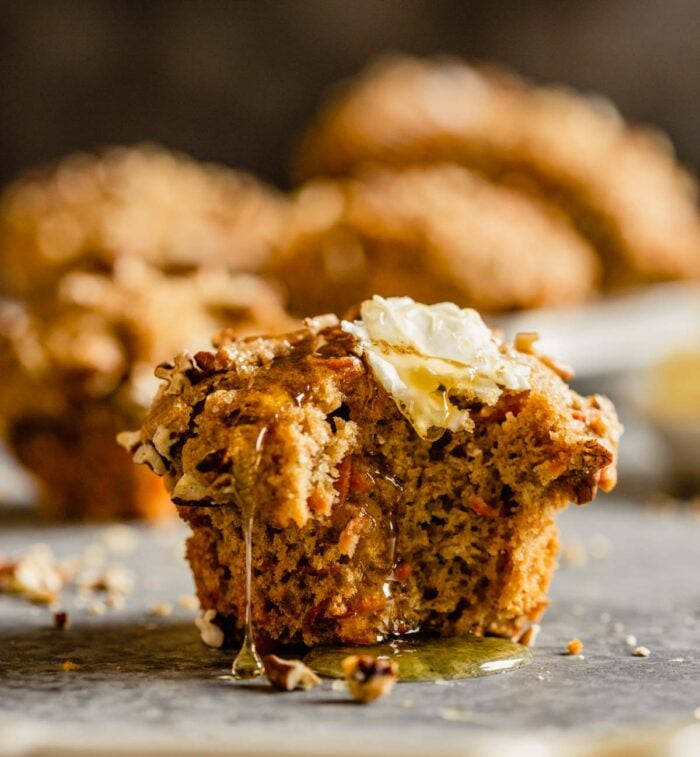 carrot muffin with a bite taken out of it set on a table with a pat of butter on top and honey dripping off it