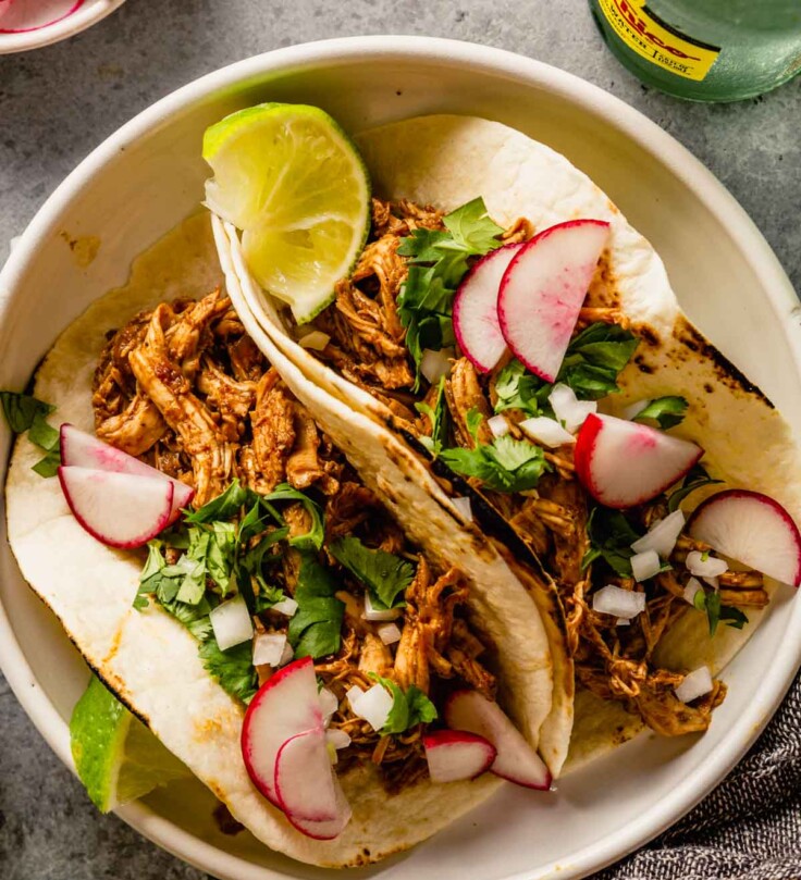 shredded chicken tacos in a white bowl topped with radish slices, lime wedges, white onion and cilantro.