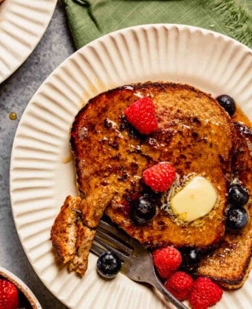 french toast topped with berries, butter and maple syrup on a white plate with a green napkin set next to it