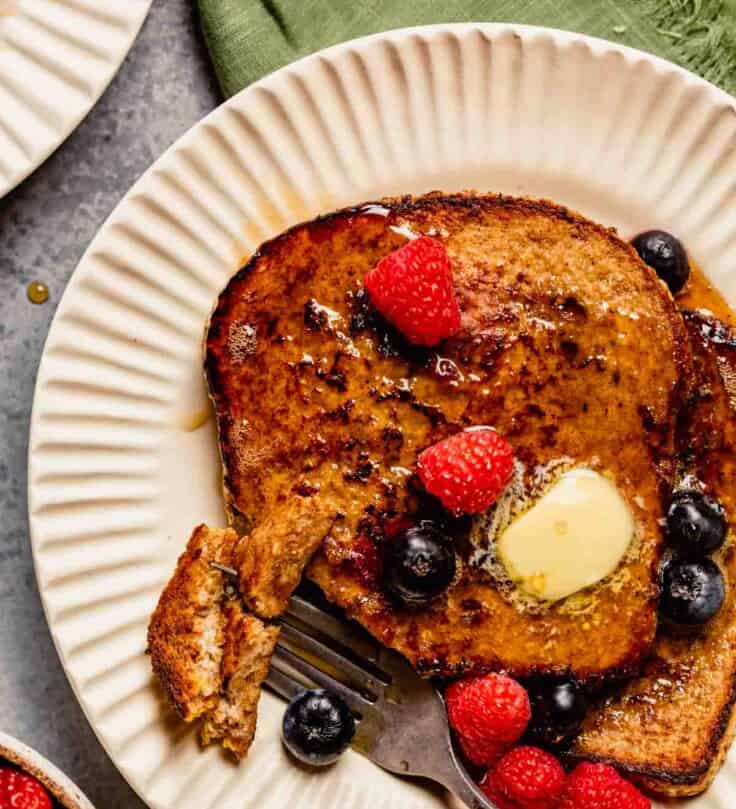 french toast topped with berries, butter and maple syrup on a white plate with a green napkin set next to it