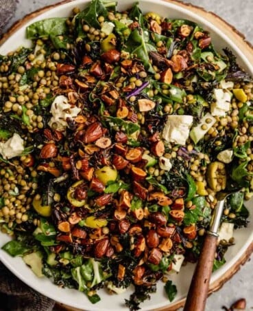 kale and lentil salad in a white bowl set on a wood board topped with feta cheese and toasted almonds