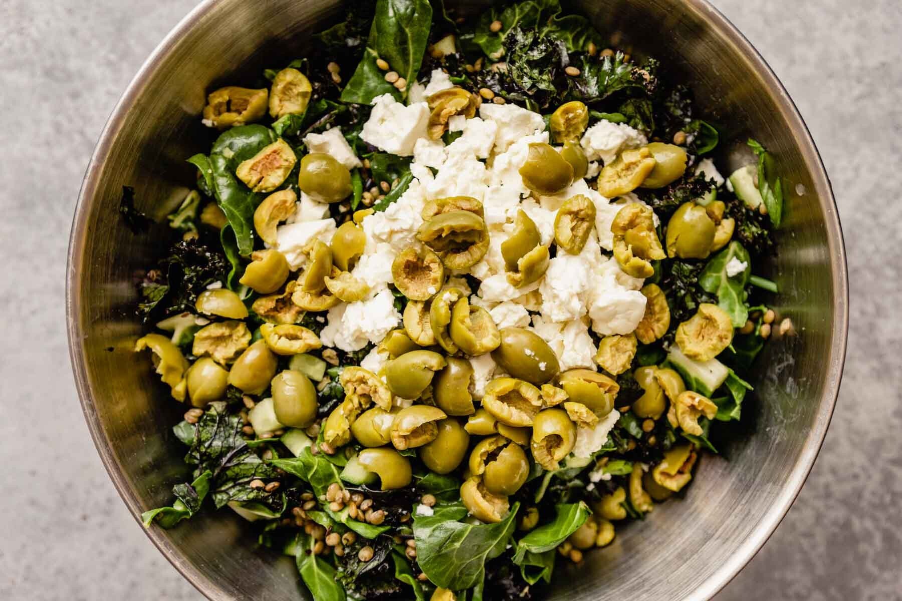 Large mixing bowl filled with swiss chard and kale topped with cooked lentils, olives and crumbled feta. 