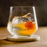 rocks glass filled with a clear liquid, orange peel and large chunk of ice set on a white coaster.