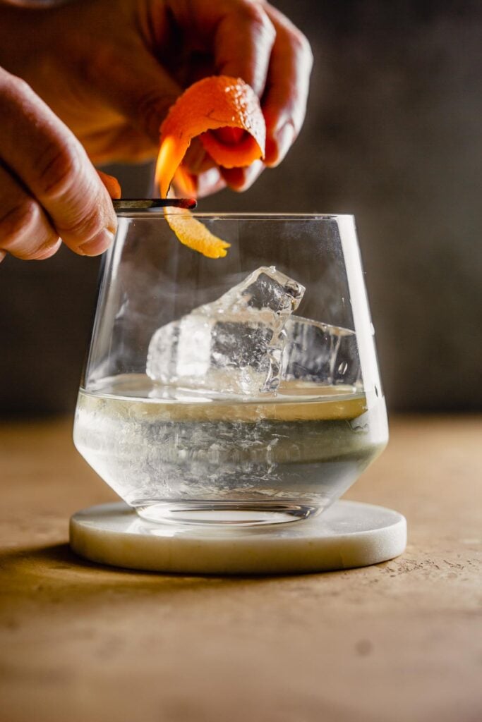 rocks glass filled with a clear liquid and large chunk of ice set on a white coaster. Someone flaming an orange peel over top