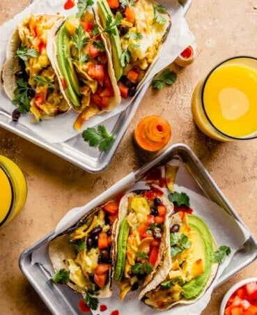 breakfast tacos filled with scrambled eggs, avocado slices, tomatoes, black beans on silver trays with parchment paper. Orange juice and hot sauce set around