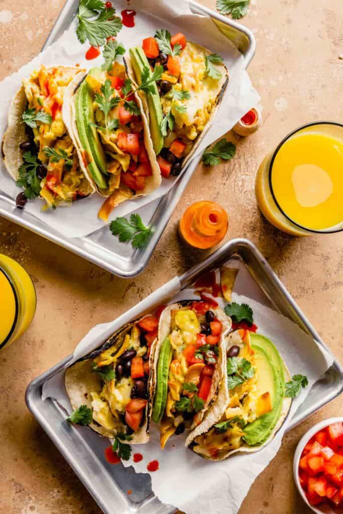 breakfast tacos filled with scrambled eggs, avocado slices, tomatoes, black beans on silver trays with parchment paper. Orange juice and hot sauce set around