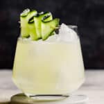 rocks glass filled with a light yellow-colored drink and topped with a cucumber ribbon