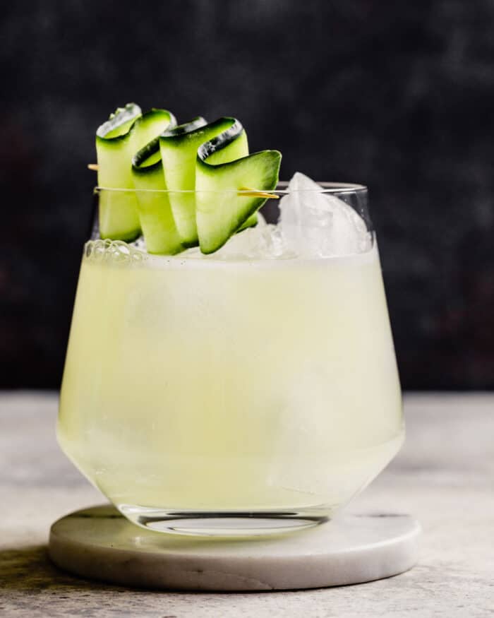rocks glass filled with a light yellow-colored drink and topped with a cucumber ribbon