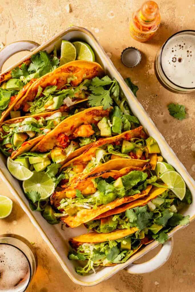 Tacos filled with an orange-colored filling and topped with shredded lettuce, avocado and lime wedges arranged in a baking dish