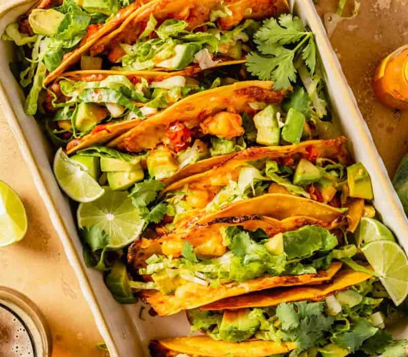 Tacos filled with an orange-colored filling and topped with shredded lettuce, avocado and lime wedges arranged in a baking dish