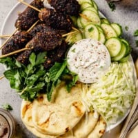 large white platter full of ground lamb skewers, shredded cabbage, sliced cucumber, herbs, and naan bread