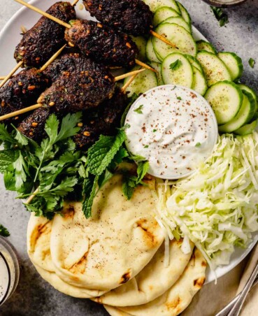 large white platter full of ground lamb skewers, shredded cabbage, sliced cucumber, herbs, and naan bread
